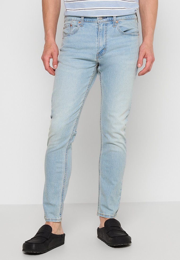 Homme portant un jean slim bleu clair et des chaussures noires à enfiler, se tenant contre un fond gris clair uni.