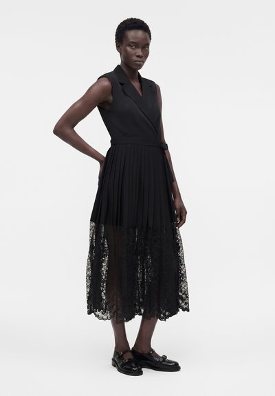 Γυναίκα με κοντά σγουρά μαλλιά που φοράει μαύρο αμάνικο φόρεμα με πλισέ φούστα και διάφανο δαντελένιο τελείωμα, συνδυασμένο με μαύρα παπούτσια με αγκράφες.