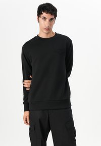 Schwarzes Sweatshirt mit Rundhalsausschnitt, langen Ärmeln und gerippten Bündchen. Besitzt ein dezentes geprägtes Logo auf der Brust. Weicher Stoff.