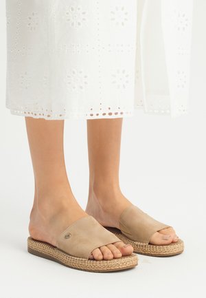 A piedi nudi in sandali scivolati in suede beige con suola in juta intrecciata. I sandali presentano una fascia larga e un design minimalista. Vestito bianco ricamato visibile.