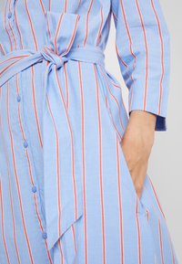 Jasnoniebieska sukienka koszulowa w pionowe czerwone paski, z długimi rękawami, zapinana na guziki z paskiem w talii i kieszeniami po bokach.