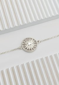 Silver armband med en rund, strukturerad berlock med strålande linjer och en liten central sten. Kedjelänkarna är fina och delikata.