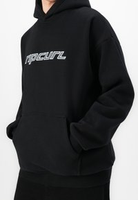 Personne portant un sweat à capuche Rip Curl noir avec poche avant, les mains dans les poches, et un logo blanc sur la poitrine sur un fond uni.