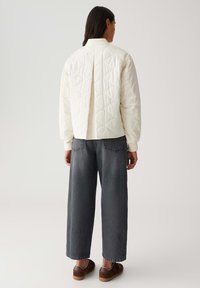 Veste matelassée de couleur crème avec un pli au dos, associée à un jean ample gris foncé et des baskets marron, vue de dos.