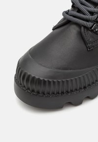 Gros plan sur l'avant d'une botte en cuir noir avec une semelle en caoutchouc texturée robuste et des lacets ronds gris-noir sur un fond clair.