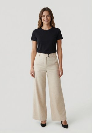 Mujer de pie, con una blusa negra de manga corta, pantalones beige de pierna ancha con doble botón y zapatos de tacón negros con punta afilada, sonriendo a la cámara.