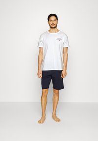 Hvit bomulls t-skjorte med "Tommy Hilfiger"-logo, sammen med marineblå shorts. Enkelt design, korte ermer og avslappet passform. Ingen synlige mønstre.