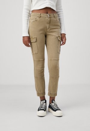 Pantalones cargo color caqui con dobladillos ajustados, bolsillos laterales y detalles de costura. Llevados con zapatillas de deporte negras y blancas. Tejido de denim texturizado.