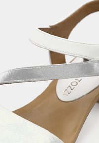 Λευκό σανδάλι με λουράκια, με γυαλιστερό ασημί λουράκι, λεία υφή και ανοιχτό καφέ πάτο. Το όνομα της μάρκας είναι ορατό μέσα στο παπούτσι.