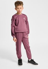 Lilla træningsdragt lavet af glat stof, der består af en sweatshirt med rund hals og taperede bukser med kontrasterende sidelænder. Sorte sneakers.