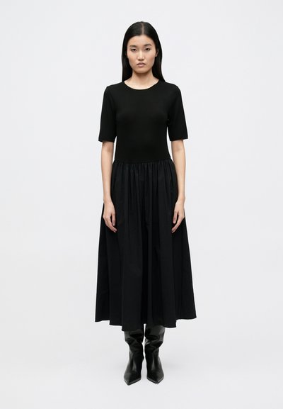 Schwarzes Kleid mit figurbetontem kurzärmeligem Oberteil und ausgestelltem, gerafftem Rock. Kombiniert mit kniehohen schwarzen spitz zulaufenden Stiefeln. Glatte Textur.