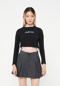Black cropped long-sleeve top with "Santa Cruz" embroidered in purple, paired with a high-waisted grey pleated skirt featuring a button.