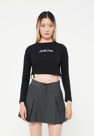 Black cropped long-sleeve top with "Santa Cruz" embroidered in purple, paired with a high-waisted grey pleated skirt featuring a button.