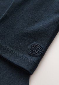 Tamsiai mėlynas medvilninis rankovės audinys su lygiu paviršiumi, šalia rankogalių siuvinėtas logotipas toniniu dizainu. Kruopščiai siuvinės kraštai.