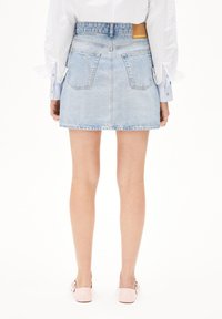 Jupe en denim bleu clair avec un ourlet effiloché, deux poches arrière et une étiquette de marque beige sur la taille, associée à des ballerines roses.