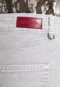 Gros plan sur la poche d'un jean en denim gris clair avec une étiquette de marque en cuir rouge MAX&Co. et un anneau métallique pour porte-clés sur la ceinture.