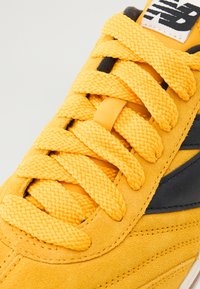 Nahaufnahme eines gelben Wildledersneakers mit geflochtenen Schnürsenkeln und schwarzen Akzenten. Der Sneaker hat eine weiche Textur und sichtbare Nähedetails.