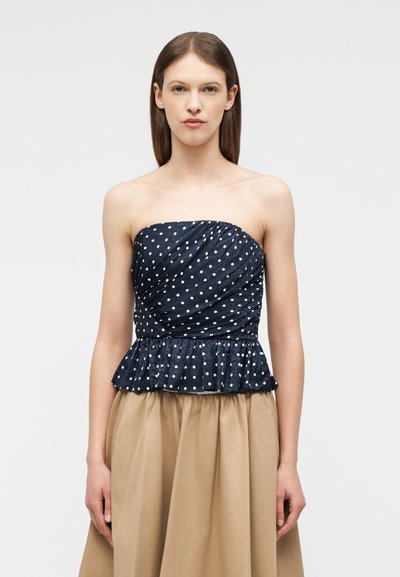 Γυναίκα με ίσια καστανή τρίχα φοράει μπλε σκούρο τοπ με πουά και βολάν στη μέση και μπεζ ψηλόμεση φούστα, στέκεται μπροστά σε λευκό φόντο.