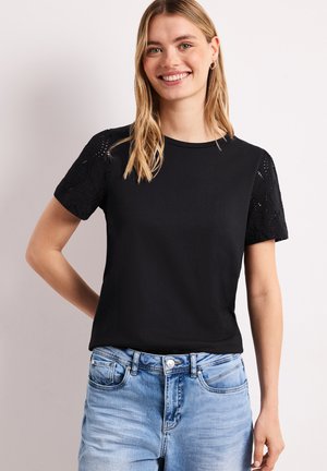 Usmívající se žena s dlouhými blond vlasy nosí černý tričko s krátkými rukávy a krajkovými rukávy a světle modré džíny, stojí na bílém pozadí.