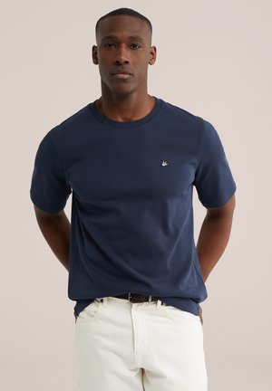 Homme portant une chemise bleu marine à manches courtes et un pantalon blanc, debout les mains derrière le dos devant un fond uni.