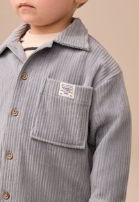 Veste chemise en velours côtelé gris avec un col, fermeture bouton et une poche à rabat avec une étiquette sur le côté gauche. Texture côtelée sur l'ensemble.