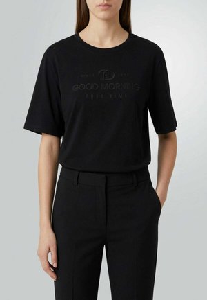 Donna con maglietta nera con la scritta "GOOD MORNING FREE TIME" e pantaloni neri sartoriali, in piedi davanti a uno sfondo grigio chiaro.