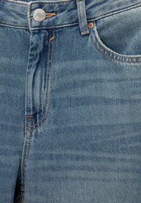 Gros plan sur un jean en denim bleu montrant le bouton, la zone de la fermeture éclair, la poche et les détails de couture orange.