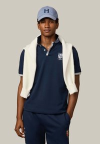 Jonge man die een marineblauw poloshirt en bijpassende broek draagt, met een lichtblauwe pet en een wit vest over zijn schouders gelegd.