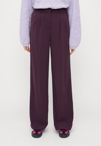 Vide ben bukser i mørk lilla stof, med fork shouldrest pleats og høj talje-design, parret med sorte sneakers med lyserøde accenter.