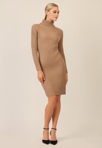Ribbet turtleneck kjole i lys brun, tætsiddende med lange ærmer, knælængde, parret med sorte stiletto hæle. Glat tekstur.