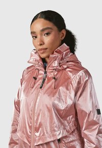 Šviesiai rožinė, blizganti, su gobtuvu striukė su užtrauktuku priekyje, raišteliais kaklo srityje ir sutrumpintu siluetu. Tekstūruotas audinys su šiek tiek blizgesio.