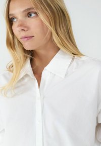 Weißes Hemd mit klassischem Kragen, das eine glatte Textur und ein minimales Design aufweist, unterstrichen durch subtile Stichdetails.