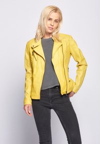 Geel leren motorjack met een aansluitend ontwerp, voorzien van een diagonale rits, textuur op de schouders en een gladde afwerking. G gedragen met een grijze top.