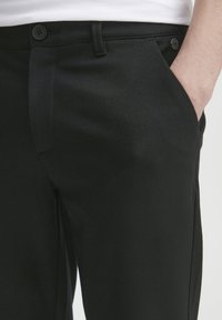 Schwarze Maßhosen aus einem glatten Stoff mit vorderen Taschen und Knopfverschluss, im klassischen geraden Schnitt.