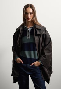 Veste oversized marron avec un col montant, superposée à un pull rayé bleu marine, vert et gris, associée à un jean bleu.