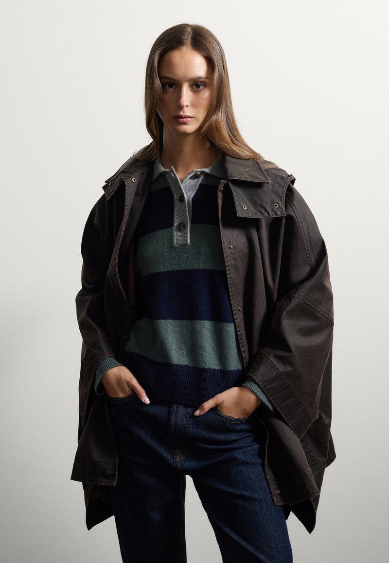 Veste oversized marron avec un col montant, superposée à un pull rayé bleu marine, vert et gris, associée à un jean bleu.