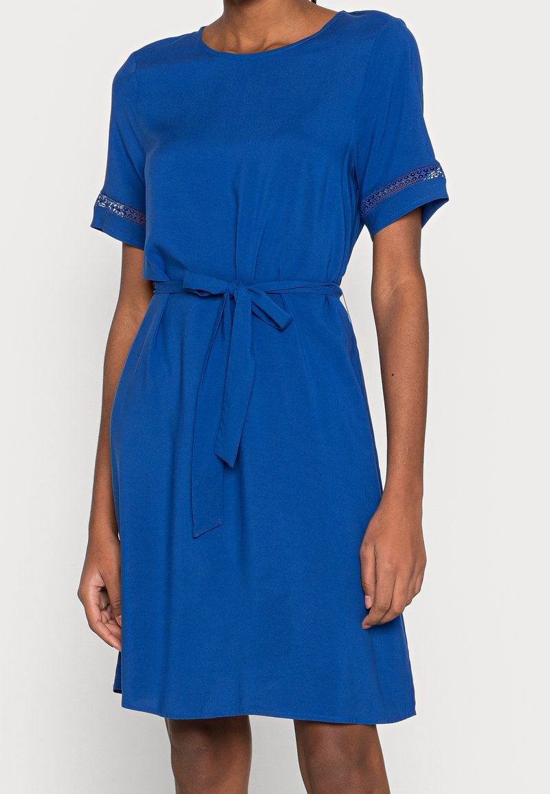 Robe bleue longueur genou avec manches courtes, ceinture en tissu à la taille et bordure décorative sur les poignets des manches portée par une personne à la peau moyennement foncée.