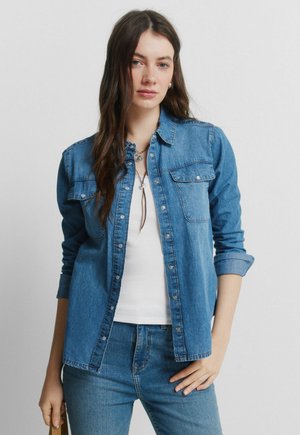 Pitkät ruskeat hiukset omaava nainen pukeutuneena valkoisen paidan päälle käännetyillä hihoilla olevaan farkkupaitaan ja farkkuihin, seisomassa yksinkertaisen harmaan taustan edessä.