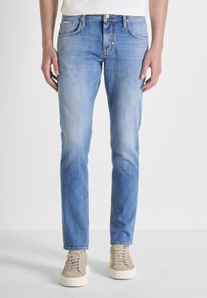 Uomo che indossa jeans slim fit azzurri chiaro e sneaker beige, in piedi con le mani rilassate lungo i fianchi davanti a uno sfondo semplice.