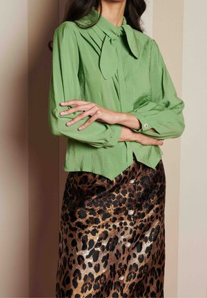 Kobieta ubrana w zieloną satynową bluzkę z wiązanym kołnierzem i długimi rękawami w połączeniu z połyskującą spódnicą na guziki w panterkę, ramiona skrzyżowane.