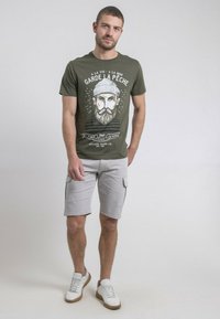 T-shirt vert olive avec un imprimé graphique d'un homme barbu, associé à un short cargo gris clair et des baskets blanches avec des accents marron.