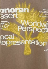 Hnědé tričko s žlutou grafikou palmy a bílým textem "Sonorská poušť," "Celosvětová perspektiva" a "Místní zastoupení."