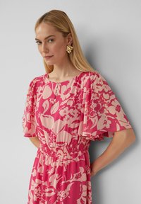 Robe fleurie en rose et rose clair, avec des manches courtes en volants, une taille smockée et des boutons au niveau du col. Boucles d'oreilles en or texturé.
