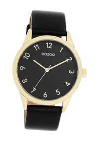 Montre analogique noire avec un boîtier rond doré, un cadran noir, des index numérotés blancs et un bracelet en cuir noir.