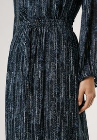 Abito lungo blu scuro caratterizzato da un tessuto testurizzato con accenti metallici, motivi a righe verticali e una vita elasticizzata con cordoncino.
