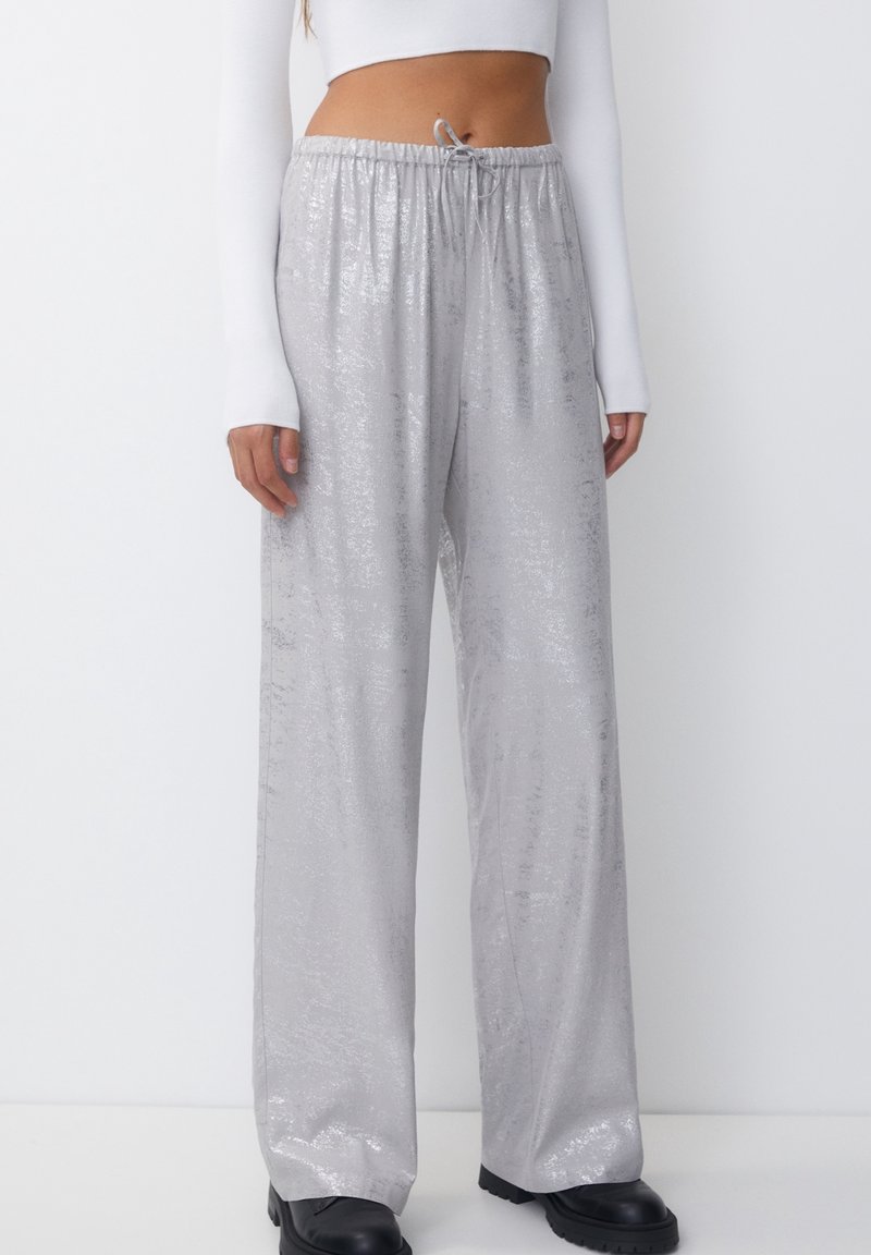 Zilveren wijd beenpantalons met een glanzende textuur en elastische tailleband. Bevat een koord en subtiele patronen. Gepaard met zwarte platformlaarzen.