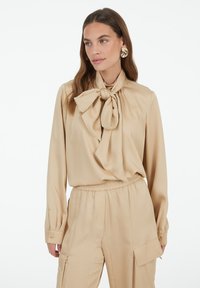 Beige blouse with a large bow tie at the neckline, long sleeves, and cuffs with button details, paired with matching beige pants. Smooth fabric texture.