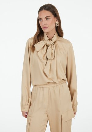 Beige blouse with a large bow tie at the neckline, long sleeves, and cuffs with button details, paired with matching beige pants. Smooth fabric texture.