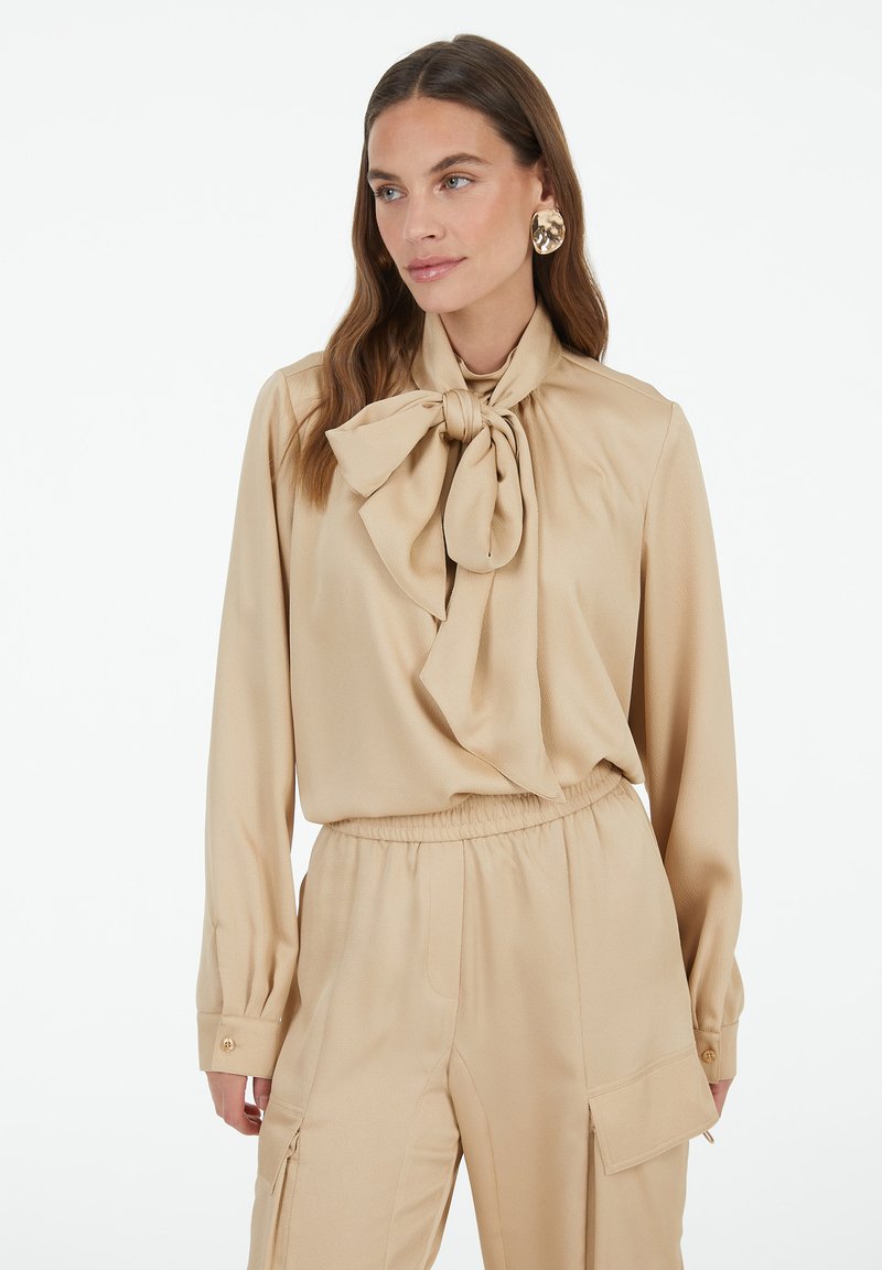 Beige blouse with a large bow tie at the neckline, long sleeves, and cuffs with button details, paired with matching beige pants. Smooth fabric texture.