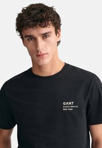 Camiseta de algodón negra con un pequeño estampado blanco que dice "GANT" y "32 EAST 39TH ST. NEW YORK" en la parte izquierda del pecho. Mangas cortas.